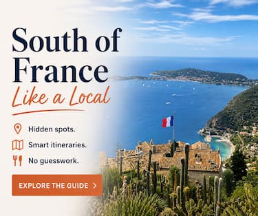 South of France travel guide by local. Panoramic view of the Côte d’Azur with terracotta rooftops, deep blue Mediterranean Sea, scattered boats, and a French flag above a hilltop village.