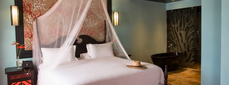 Elegant hotel room at Villa Delisle Hôtel & Spa in Saint-Pierre with canopy bed, hat on white linens, and a black standalone bathtub.