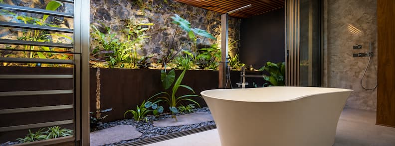 Open-air bathroom at a luxury hotel in La Réunion with a freestanding bathtub, tropical plants, and stone walls under a slatted wooden ceiling.