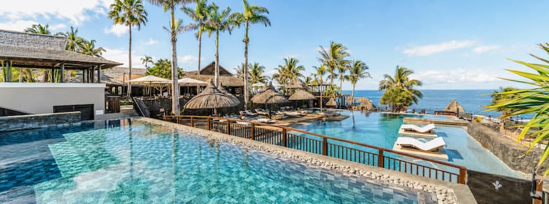 Luxury beachfront resort in La Réunion with tiered infinity pools, sun loungers, thatched umbrellas, and ocean views framed by palm trees.