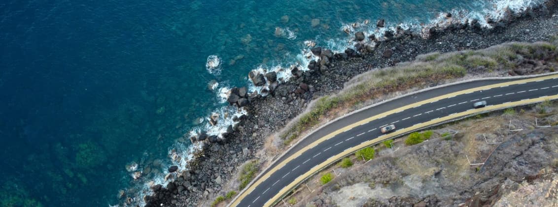 do you need a car in la reunion road trip reunion island Coastal road on La Réunion Island seen from above, with cars driving beside volcanic cliffs and the Indian Ocean shoreline.
