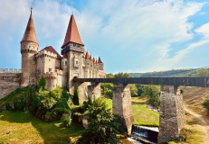 Corvin Castle in Transylvania Romania with medieval towers, stone bridge, and countryside landscape under a blue sky.