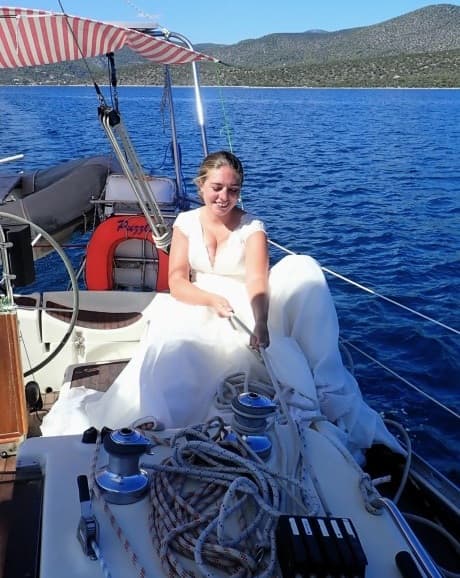 Mirela Letailleur sailing in Greece aboard her sailboat in the Mediterranean Sea, surrounded by deep blue water and rocky coastline in the Saronic Gulf.