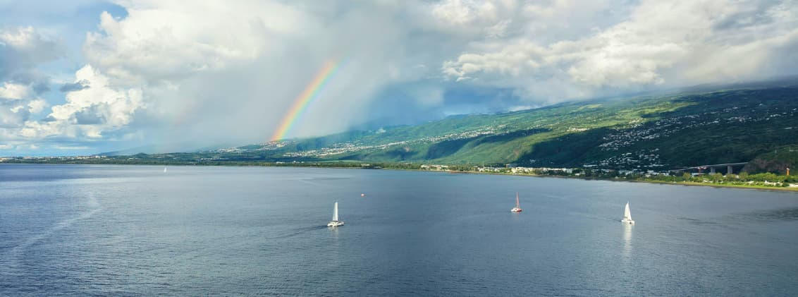 why la reunion is different indian ocean island Rainbow above the coastline of La Réunion with volcanic mountains, ocean views, and sailboats along the Indian Ocean shore.