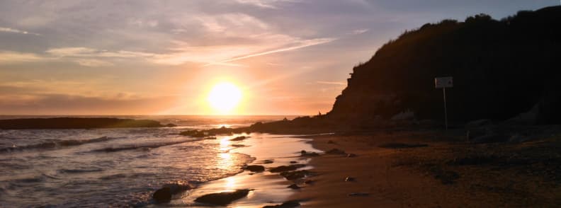 Sunset over Anzio beach near Rome, with shallow waves, wet sand, scattered rocks, and a dark coastal headland.