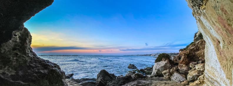 View from a rocky sea cave near Anzio beach, framing blue Tyrrhenian water, scattered boulders, and a pastel sunset along the Lazio coast.