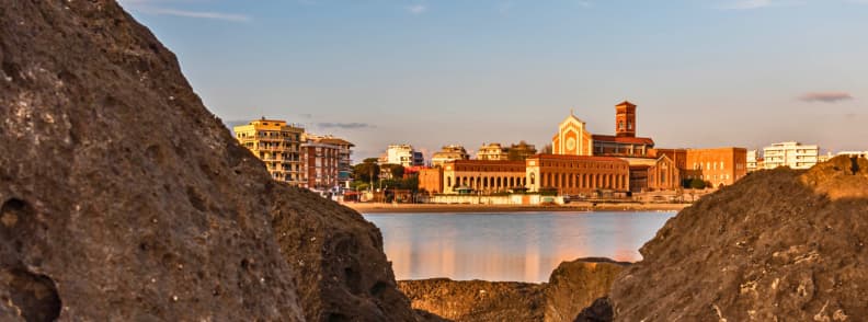 Basilica of Our Lady of Graces on Nettuno Waterfront Basilica of Our Lady of Graces and Saint Maria Goretti beside the Nettuno waterfront, viewed across calm water between coastal rocks.