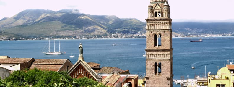Gaeta Cathedral bell tower rising above terracotta rooftops, with sailboats, a cargo ship, the Gulf of Gaeta, and mountains across the water.