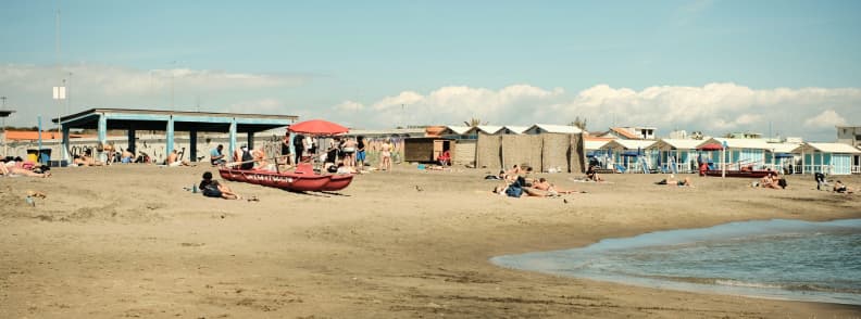 Sandy Lido di Ostia beach near Rome with swimmers, sunbathers, colourful beach cabins, lidos, and a red lifeguard rescue boat.