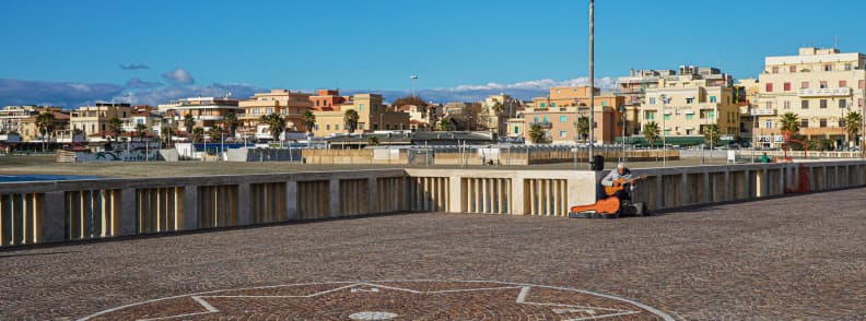 Street musician playing on the Lido di Ostia seafront promenade, with waterfront buildings, palm trees, and the Lazio coast behind him.