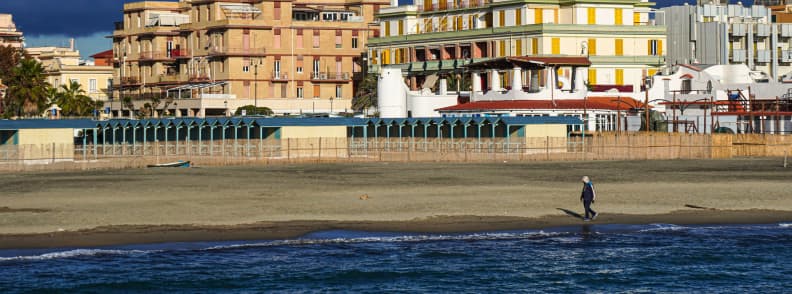 Ostia beach near Rome with a lone walker beside the water, dark sand, seafront lidos, palm trees, and urban waterfront buildings.