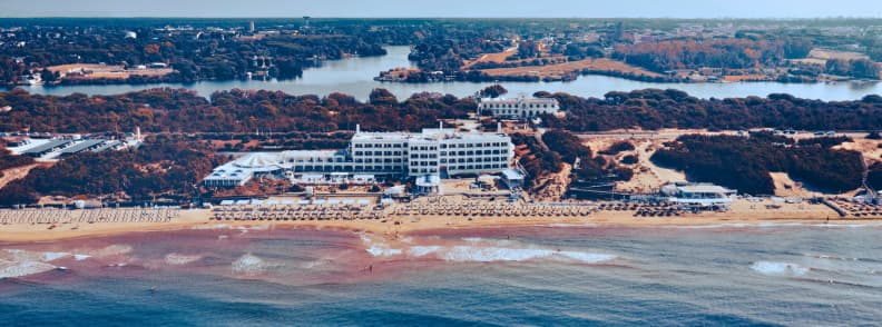 Aerial view of Sabaudia beach near Rome with a sandy shoreline, beachfront hotel, rows of umbrellas, coastal dunes, and Lake Paola inland.