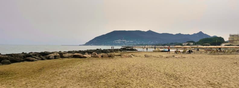 Wide sandy San Felice Circeo beach on the Lazio coast, with rock breakwaters, beach umbrellas, calm sea, and Mount Circeo in the distance.
