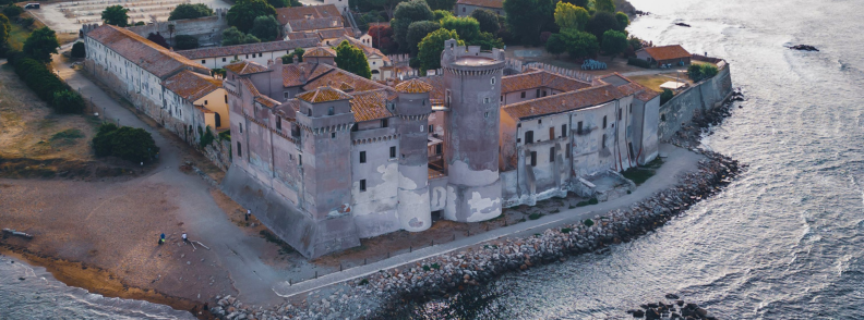 Aerial view of Santa Severa Castle beside a sandy beach and rocky Tyrrhenian shoreline near Civitavecchia cruise port.