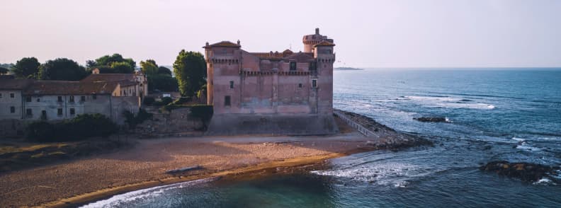 Aerial view of Santa Severa beach near Rome with the medieval seaside castle, a curved sandy cove, rocky reefs, and blue Tyrrhenian water.