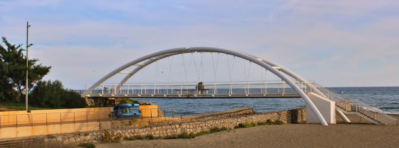White arched pedestrian bridge beside Sperlonga beach, with sandy shore, stone walls, coastal trees, and the Tyrrhenian Sea beyond.