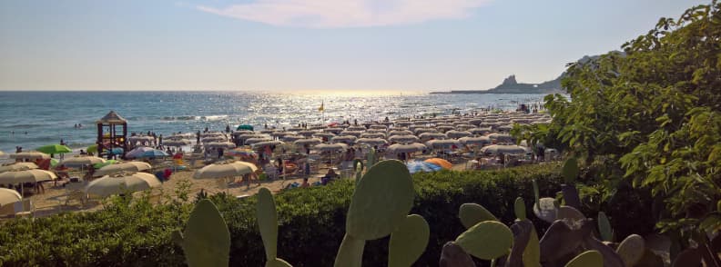 Crowded Sperlonga beach in summer with rows of lido umbrellas, swimmers, prickly pear cactus, and Torre Truglia on the distant headland.