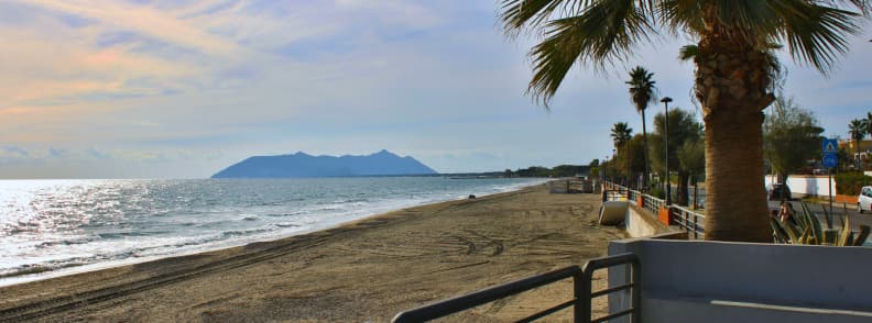Terracina beach near Rome with a broad sandy shore, palm-lined promenade, calm Tyrrhenian Sea, and the Circeo headland on the horizon.