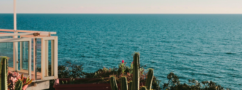 Sea-view balcony at accommodation near Anzio beach, with glass railings, potted cacti, coastal plants, and the blue Tyrrhenian Sea.