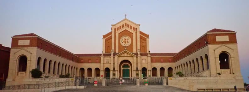 visit Basilica of Our Lady of Graces in Nettuno Italy Front view of the Basilica of Our Lady of Graces and Saint Maria Goretti in Nettuno, with brick façades, arcades, a rose window, and gated steps.