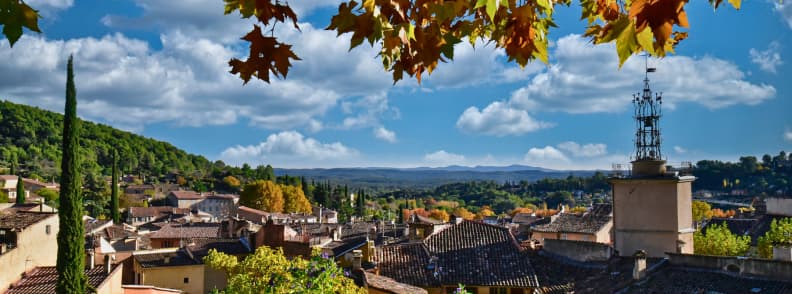 Autumn in Provence over a hill village in Cotignac with terracotta rooftops, golden plane-tree leaves, cypress trees, and distant wooded hills beneath a blue sky.