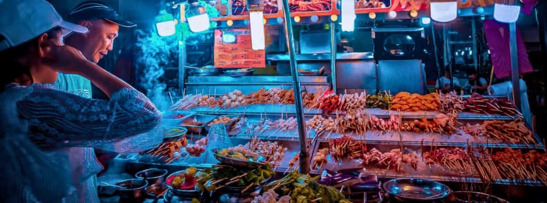 Vendor preparing grilled skewers at a brightly lit Kuala Lumpur street food stall during an evening market.