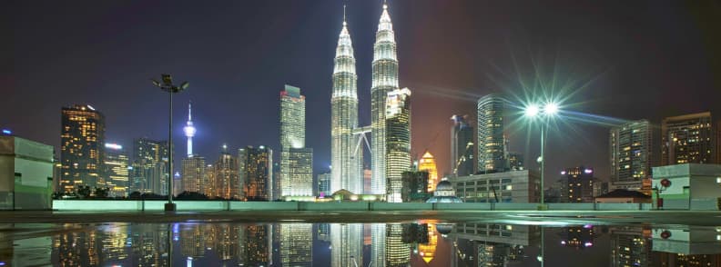 Petronas Twin Towers and KL Tower illuminated above the Kuala Lumpur skyline, with city lights reflected in a rooftop pool.