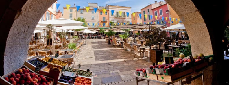 Autumn Food Markets and Harvests on the Cote d Azur Autumn food market on the Côte d’Azur, with crates of peaches, tomatoes, melons, and seasonal produce beside café terraces in a colorful town square.