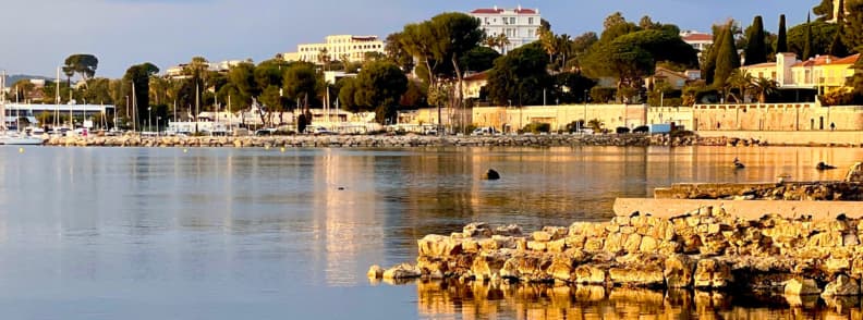 French Riviera Autumn Year Round Cities and Services Calm French Riviera waterfront in Antibes in autumn, with rocky shoreline, Mediterranean water, trees, hotels, and historic coastal buildings.