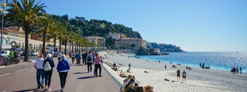 Nice in Autumn French Riviera Day One Itinerary Nice in autumn, with people walking along the Promenade des Anglais beside the pebble beach, palm trees, and bright Mediterranean water.