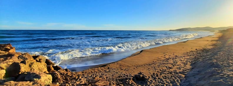Base Nature Beach Frejus Golden Hour Mediterranean Coast Mediterranean waves breaking along Base Nature beach in Fréjus, with golden sand, sunlit rocks, footprints, and distant coastal hills.
