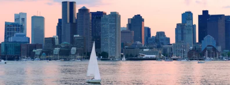 Free things to do in Boston guide. Sailboat crossing Boston Harbor with the downtown Boston skyline in the background at sunset.