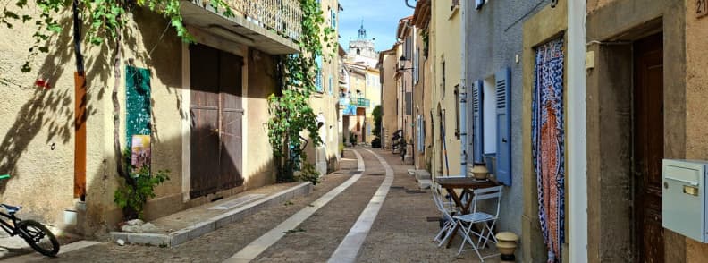 Full Day in Mons Var Old Village Streets Quiet old street in Mons Var with pastel houses, blue shutters, climbing vines, and small outdoor tables along the paved lane.