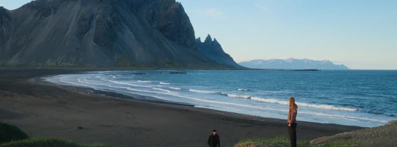 Two travelers on a black sand beach in Iceland, with steep mountains rising behind the coastline.