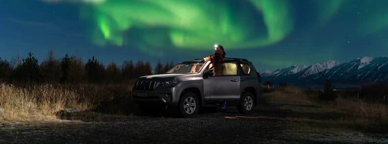Northern Lights Above a Parked SUV in Iceland Green northern lights above a parked grey SUV, with a person beside the vehicle and snowy mountains beyond.