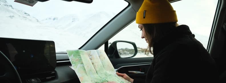 Planning an Iceland Road Trip with a Paper Map Woman wearing a yellow knit hat reading a folded map in the passenger seat of a car, with snowy mountains outside.