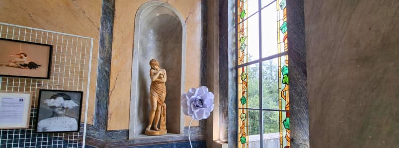 Villa Aurelienne Frejus Historic Statue and Festival Art Classical female statue in a wall niche at Villa Aurélienne in Fréjus, beside a large lavender flower sculpture, an arched stained-glass window, and photography festival displays.