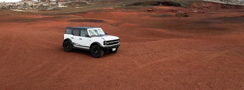 White 4x4 Vehicle on Red Volcanic Terrain White four-wheel-drive SUV parked on a broad red gravel plain with low rocky mountains in the background.
